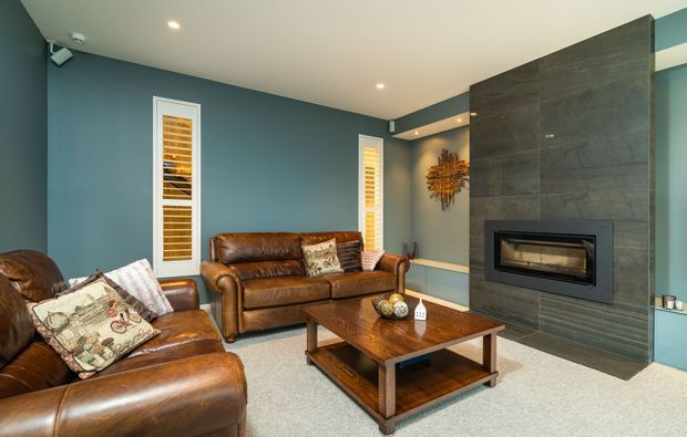 A refined living room with deep blue-grey walls, cream ceiling, pale carpet, and rich brown leather seating arranged around a substantial timber coffee table. A sleek built-in fireplace is set within a dark large-format tiled feature wall, complemented by narrow shuttered windows, recessed lighting, and a warm sculptural wall light.