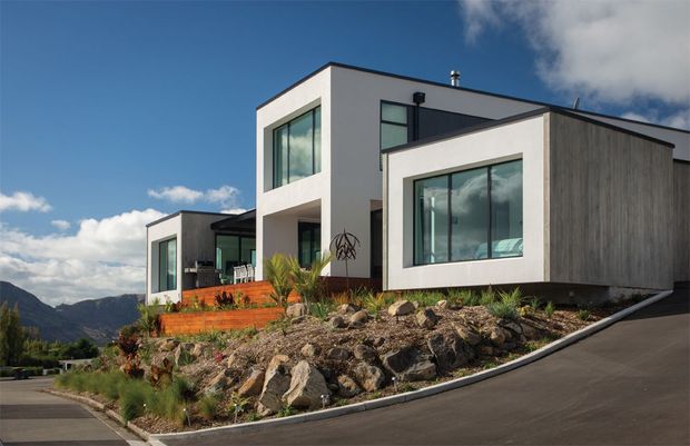 Modern multi-level residential home on a sloped, landscaped site, featuring crisp white rendered volumes, dark vertical timber-look cladding, expansive black-framed glazing, and a flat roof. The home is approached by a paved driveway and surrounded by exposed rock landscaping, ornamental grasses, and mountain views.