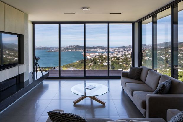 A contemporary living room with expansive floor-to-ceiling glazing overlooking a turquoise bay and densely built hillside landscape. The room features a large neutral sectional sofa, round glass coffee table with timber cross-base, dark tiled flooring, minimalist built-in cabinetry, recessed ceiling lighting, and black-framed sliding or fixed glass panels.