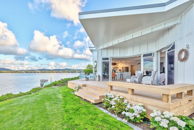 Bright waterfront home exterior with a white-clad contemporary pavilion, broad projecting roof, expansive sliding glass doors, and a timber deck with steps leading to a manicured lawn. The open interior appears to include a living area and kitchen, while the landscape overlooks calm water and distant hills. Pale coastal colours, hydrangeas, and outdoor seating create an airy, relaxed setting.
