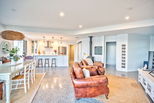 Bright open-plan interior combining a kitchen, dining area and living room. The space features polished light terrazzo-style flooring, white cabinetry and walls, a kitchen island with pendant lighting, a timber dining table, and substantial cognac leather sofas arranged around a media unit. A large woven pendant and natural-fibre rug add warm texture to the otherwise light, open room.