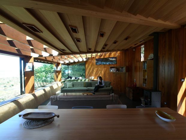 A warm, wood-lined open-plan interior viewed from a dining table toward a living area. The room features extensive timber wall and ceiling lining, exposed timber ceiling beams, recessed spotlights, built-in shelving and cabinetry, a large sectional sofa, and floor-to-ceiling glazing with expansive landscape views. A long dining table occupies the foreground, while the living room is the dominant space beyond.
