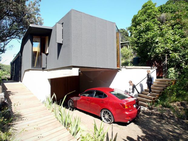 Exterior view of a contemporary multi-level house set into a sloped, landscaped site. The home has a strongly geometric dark grey box-like upper volume, large timber-framed glazing with projecting shutters, white rendered lower walls, and an integrated undercroft driveway/garage area. Timber stairs and a boardwalk connect the driveway to the garden, with mature vegetation surrounding the building.