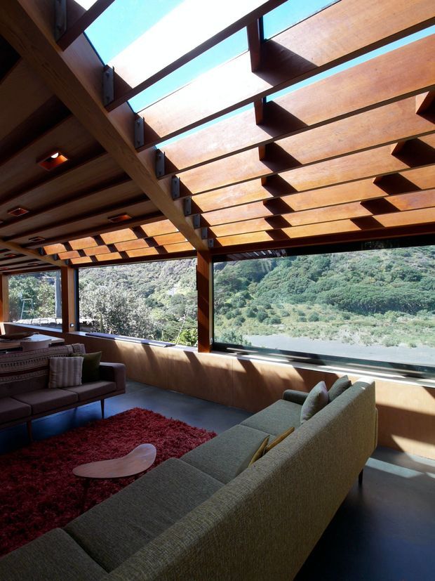A dramatic living room with expansive glazing overlooking a lush valley and river, framed by a heavily timbered ceiling with repeating projecting wood sun-shading fins and skylight openings. The room contains large contemporary sofas, a red shag rug, and a low sculptural coffee table. Strong sunlight creates warm timber tones and high-contrast shadows throughout the space.