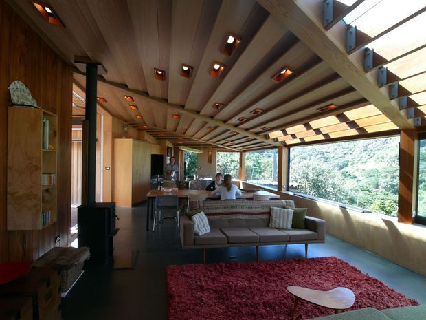 A warm, timber-lined open-plan living space with a lounge area, compact kitchen and dining zone. The room features a dramatic exposed timber beam and slatted ceiling with recessed lighting, extensive corner glazing, built-in timber cabinetry, a dark floor, and a red area rug. Lush vegetation is visible outside through the windows.