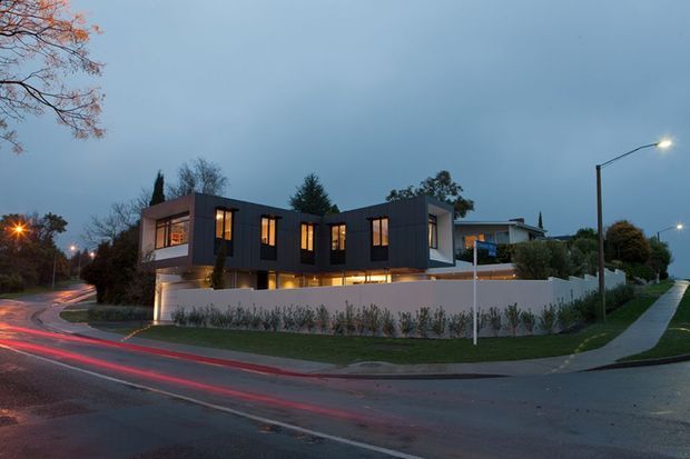 Twilight street view of a contemporary multi-storey residence on a landscaped suburban corner site. The dark-clad upper volume has large illuminated windows and projects over a lighter lower level, enclosed by a white rendered boundary wall. Wet roads, street lighting, a sidewalk, and mature planting frame the modern house.