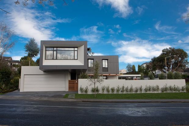 Street-facing view of a contemporary multi-level suburban residence with a strong rectilinear composition. The house features a projecting upper-level volume, dark grey rendered and panelled exterior walls, broad glazing, a large integrated garage, timber entry screening, a low white boundary wall, and restrained front landscaping.