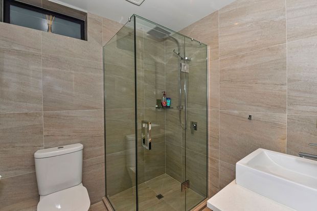 A contemporary bathroom with large beige stone-look wall and floor tiles, a frameless glass corner shower enclosure with chrome fixtures, a white toilet, and a white integrated vanity basin. A small high-level window provides natural light.