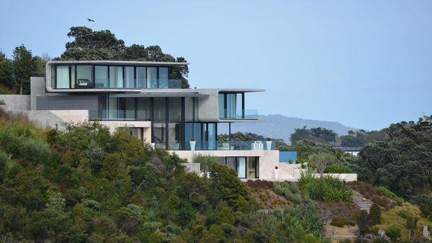 A contemporary multi-level house set into a heavily vegetated sloping site. The building features strong horizontal cantilevered forms, extensive floor-to-ceiling glazing, pale concrete or rendered walls, and flat roofs. A distant coastal or hilly landscape is visible beyond the property.