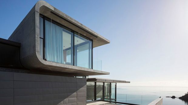 Exterior architectural view of a modern coastal residence with strong cantilevered rectangular forms, exposed concrete construction, dark horizontal wall cladding, and expansive floor-to-ceiling glazing. The building projects toward a calm ocean horizon, with a lower glazed level and roof terraces visible. The composition emphasizes crisp contemporary geometry and the waterfront setting.