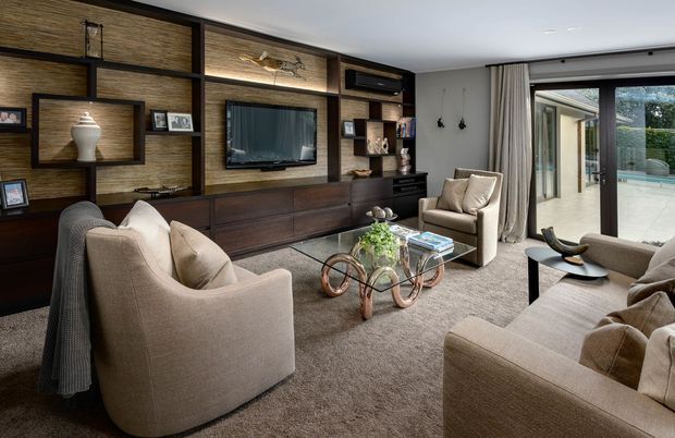 Refined contemporary living room with a large dark timber feature wall and integrated entertainment cabinetry, neutral upholstered lounge furniture, a glass coffee table with sculptural metal legs, and full-height curtains opening to a bright outdoor terrace and pool area.