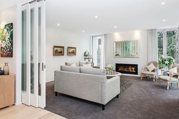 Bright, refined living room with a large light-grey sectional sofa, patterned rug, fireplace, framed artwork, and woven accent chairs. Full-height glazed folding doors open toward an adjoining space, while large windows and pale walls create an airy contemporary setting.