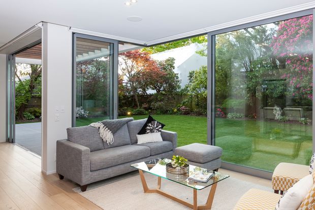 Bright contemporary living room with a grey sectional sofa, glass coffee table, light timber flooring, and extensive floor-to-ceiling glazing opening views toward a landscaped garden and covered outdoor area. The room has a clean, airy indoor-outdoor connection with restrained neutral furnishings and white walls.