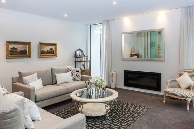 A refined, bright living room with a neutral upholstered sofa arrangement, ornate glass-topped coffee table, patterned area rug, framed artwork, striped accent chairs, full-height curtains, and a recessed black fireplace beneath a large framed mirror.