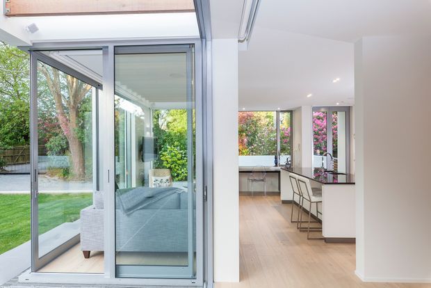 Bright contemporary open-plan interior viewed through large aluminium-framed sliding and folding glass doors. The image shows a light timber-floored living area with a pale sofa, opening into a white kitchen with a dark benchtop and bar stools. Extensive glazing connects the interior to a landscaped garden, creating an airy indoor-outdoor connection.