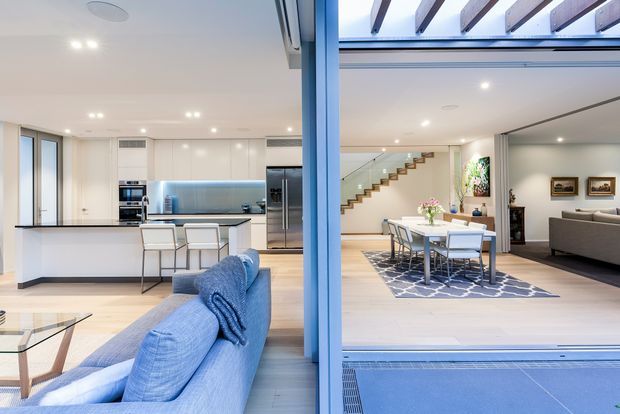Bright open-plan contemporary home interior viewed through large glazed openings, showing a white kitchen with island and integrated appliances on the left, a blue sofa in the foreground, and a dining and living area on the right. The space features pale timber flooring, extensive glazing, recessed lighting, a timber open-riser staircase, and an indoor-outdoor connection with a covered pergola and blue-toned exterior surface.