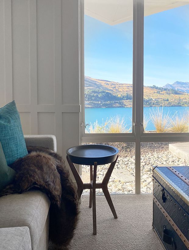 A bright, cozy sitting area beside full-height windows overlooking a vivid blue lake and surrounding sunlit mountains. The interior features a light upholstered armchair with a teal cushion and fur throw, a small dark round side table, a pale carpet, and a decorative trunk. The wall has simple recessed panel detailing, while the glazing provides a strong connection to the scenic landscape.