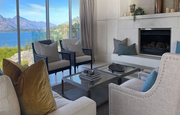 Refined living room overlooking a lake and mountain landscape through full-height glazing. The room features a white paneled fireplace wall with a dark inset firebox, neutral upholstered seating, metallic-framed glass coffee tables, layered cushions, and soft curtains. The arrangement is bright, elegant, and inviting, with strong indoor-outdoor visual connection.