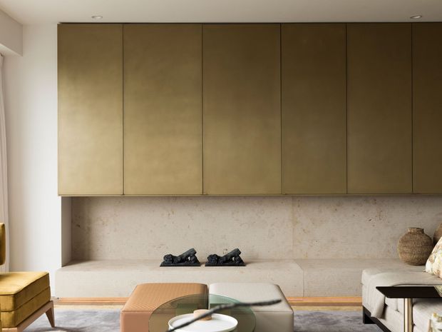 Warm, refined contemporary living room with a full-width built-in bronze-toned cabinetry wall, textured pale stone or plaster feature wall, and low sculptural seating around a glass-topped coffee table. The room has soft neutral finishes, recessed ceiling lighting, and understated natural decor.