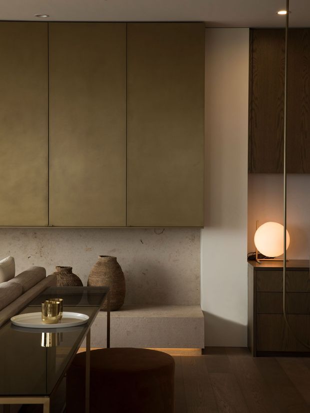 A warm, moody contemporary living room with integrated dark taupe cabinetry, a textured pale stone or plaster feature wall, built-in bench seating, and dark timber flooring. A low glass coffee table, neutral sofa, woven vessels, and a glowing spherical lamp create a refined, softly lit interior.