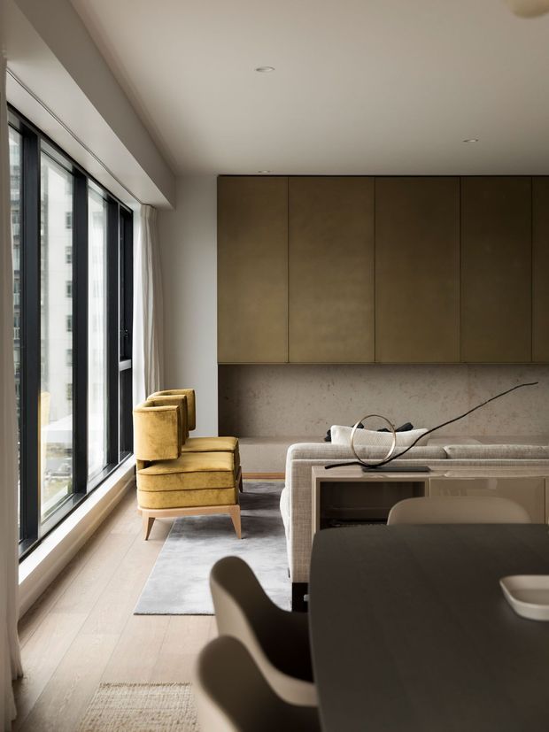 A refined open-plan apartment interior with a living and dining area. Full-height black-framed glazing runs along one side, opening the room to abundant natural light. The space features a warm mustard lounge chair, a pale upholstered sofa, a dark dining table with curved chairs, light timber flooring, a large soft grey rug, and a full-width bronze-brown built-in wall cabinet above a mottled stone or concrete-effect panel.