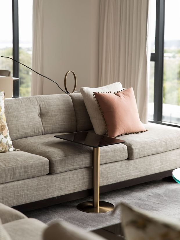 A close-up of a refined contemporary living room featuring a large textured light-grey sectional sofa, blush and cream accent cushions, a slim dark side table with a brass-gold pedestal, floor-to-ceiling windows, and full-length neutral curtains. The room has a soft, warm-neutral palette with elegant modern furnishings and abundant natural light.