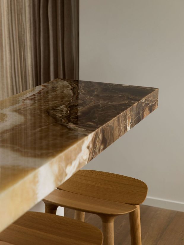 Close-up interior view of a sculptural dark brown stone or stone-look benchtop with dramatic natural veining and a thick waterfall-style edge, paired with warm timber stools. Floor-to-ceiling brown curtains, a neutral wall, and timber flooring create a refined, warm setting.