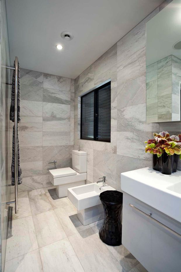 A bright, compact contemporary bathroom clad in large-format pale grey marble-look tiles across the floor and walls. The room includes a frameless glass shower with a vertical heated towel rail, wall-hung toilet, freestanding white basin, white floating vanity with chrome hardware, black-framed window, large mirror, recessed ceiling lights, and a black textured waste bin. A small floral arrangement adds colour on the vanity.