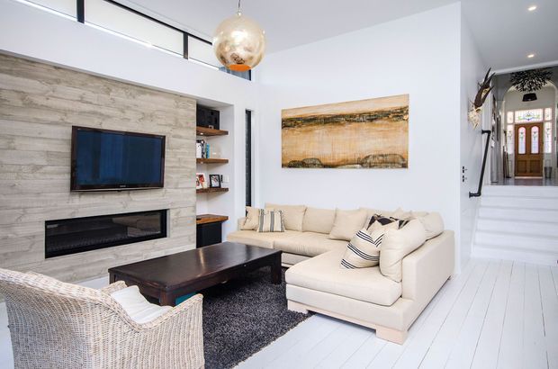 Bright, contemporary living room with white-painted timber flooring, a pale stone or concrete-look feature wall incorporating a wall-mounted television and linear fireplace, built-in open shelving, cream sectional seating, a dark coffee table, textured rug, and a woven armchair. A sculptural pendant light hangs above the seating area, while steps lead to a hall and glazed entry door in the background.