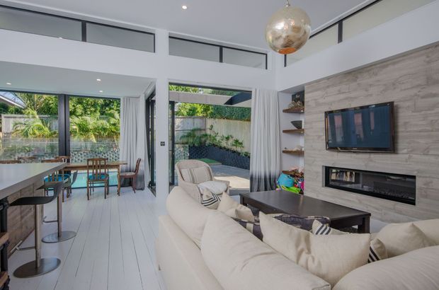 Bright open-plan living and dining area with a contemporary coastal feel. A large cream sectional sofa faces a wall-mounted television and linear fireplace set into a light timber-look feature wall. The space has white-painted timber flooring, a dining table and bar seating, high clerestory glazing, sheer curtains, and large glazed doors opening to a landscaped outdoor courtyard or patio.