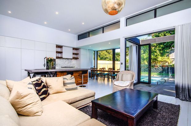 Bright open-plan contemporary interior combining a living room, kitchen and dining area. The space features a large cream sectional sofa, dark timber coffee table, white full-height cabinetry, a marble-look kitchen splashback, timber island cabinetry, extensive black-framed glazing and a glazed opening to a landscaped outdoor area. High-level clerestory windows and recessed ceiling lights contribute to the airy, spacious feel.