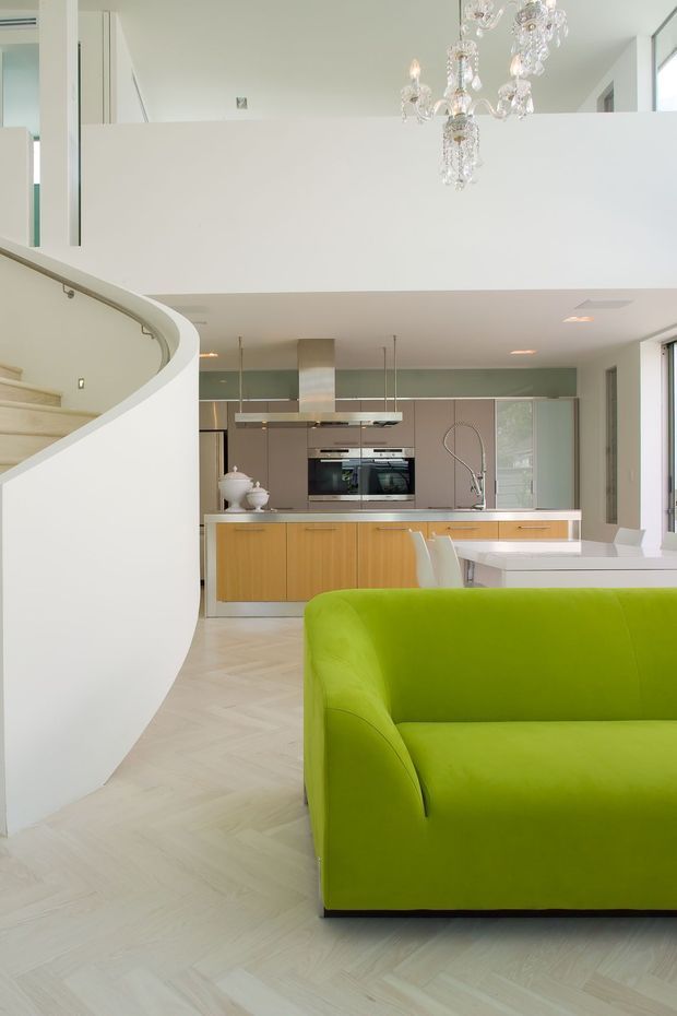 Bright open-plan contemporary interior featuring a white kitchen with timber cabinetry, integrated appliances, a pale stone island, and a white dining table. A vivid lime-green sofa occupies the foreground, while a sweeping curved staircase and a suspended crystal chandelier add sculptural and luxurious details. The pale herringbone timber floor and double-height volume create an airy, spacious feel.