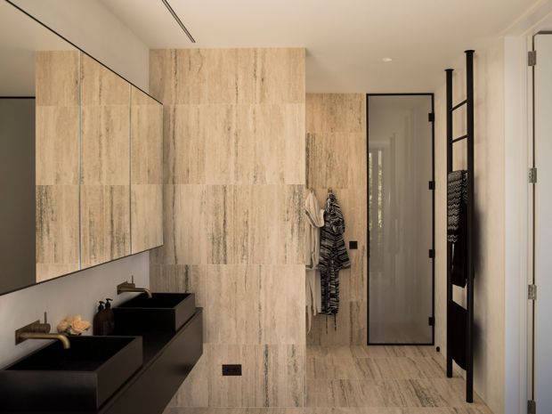 Contemporary bathroom with a double black vanity and integrated basins beneath a large wall mirror. The walls and floor are clad in large-format beige stone-look tiles with pronounced linear veining. A frameless glass shower enclosure with black hardware is visible at the rear, alongside a black heated towel rail and wall-mounted accessories. The space has clean lines, recessed lighting, and a neutral, refined palette.