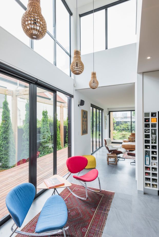 Bright contemporary open-plan living area with a dramatic double-height void, floor-to-ceiling glazing, and a series of woven pendant lights. The room features grey tiled flooring, white walls, dark-framed sliding glass doors, colourful modern lounge chairs, a patterned rug, and a dining or sitting area extending toward a landscaped garden. A tall built-in display shelf and framed artwork add visual interest.