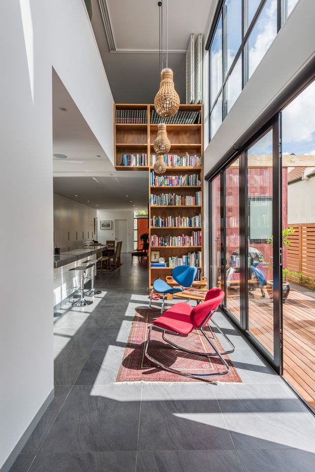A bright, double-height contemporary interior combining a kitchen and library/lounge zone. Full-height black-framed glazing opens onto a timber deck, while a tall built-in timber bookshelf rises to the ceiling. A stone kitchen island with stools sits to the left, and colourful lounge chairs rest on a patterned rug beneath sculptural woven pendant lights. Large grey floor tiles and white walls create a clean, sunlit backdrop.