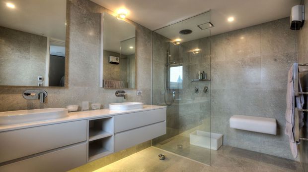 Contemporary bathroom with a double vanity, large frameless glass walk-in shower, wall-hung toilet, and full-height grey stone-look wall and floor finishes. The room has warm recessed and under-vanity lighting, mirrored cabinetry, and clean-lined built-in storage.