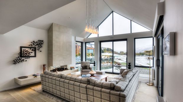 A bright, spacious contemporary living room with a large modular sectional sofa arranged around a low coffee table and a fireplace feature wall. The room has a dramatic pitched ceiling, pendant chandelier, extensive floor-to-ceiling glazing, and views toward a snowy landscape and water beyond. Neutral finishes, light timber flooring, and restrained decor create an airy, refined atmosphere.