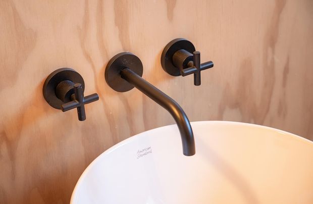 Close-up of a wall-mounted matte dark metal bathroom faucet with separate cross-handle controls above a white freestanding basin. The fixtures are mounted against a warm natural timber veneer wall, creating a refined, minimalist bathroom detail.