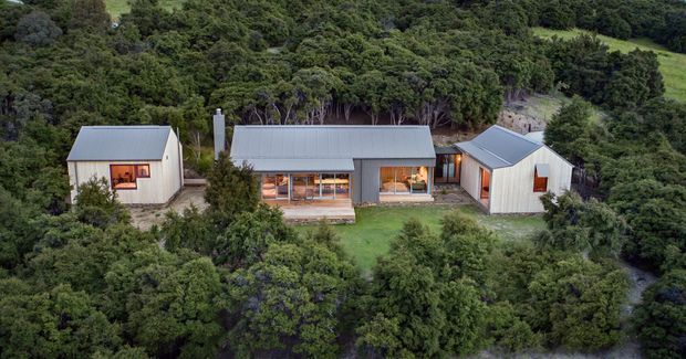 Aerial view of a rural single-storey residential compound composed of three simple gabled buildings arranged around a central open lawn and deck. The buildings have corrugated metal roofs, light-colored vertical cladding, and warm-lit windows, surrounded by dense native bush on a sloping site.