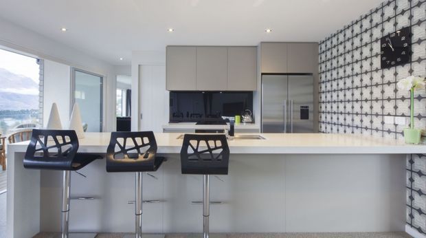 Bright contemporary kitchen with a long white island and three black bar stools, sleek light cabinetry, stainless-steel double-door refrigerator, dark benchtop work zone, and a striking black-and-white geometric tiled feature wall. Large glazing at left provides views toward a mountainous landscape. The space feels clean, open, and modern with strong graphic contrast.