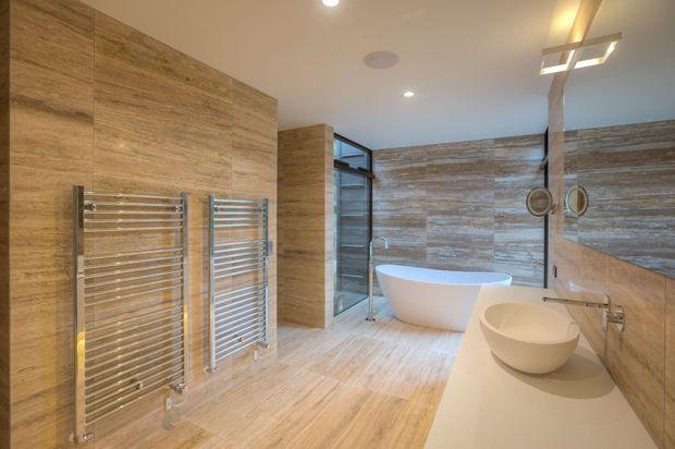 Contemporary luxury bathroom featuring extensive warm beige stone-look wall and floor finishes, a freestanding white bathtub, double vessel basins on a long vanity, large frameless glazing, and chrome heated towel rails. Recessed ceiling lighting and a sculptural pendant add refined detail.