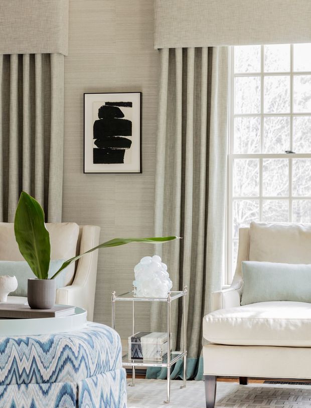 A refined sitting area with layered neutral linen curtains, textured wall covering, and a large multi-pane window. Cream upholstered seating surrounds a small glass-and-metal side table, while a blue-and-white patterned ottoman, potted tropical plant, and bold black abstract artwork add contrast. The room feels bright, calm, and tailored.