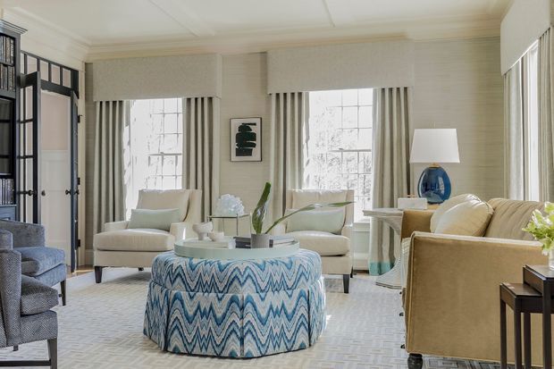 Bright, refined living room with two large windows, layered neutral furnishings, upholstered armchairs and sofa, a patterned round ottoman, framed artwork, and a blue glass lamp. The room features traditional architectural detailing with panelled walls, ceiling moulding, tall drapery, and dark-framed glazed cabinetry or doors at the left.