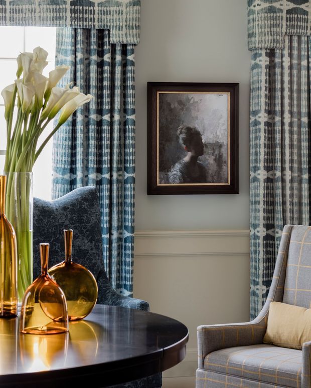 A refined sitting room vignette with blue patterned full-length curtains, a dark polished round table, amber glass vases, a patterned upholstered armchair, a framed portrait, and tall white flowers. The room has pale grey-green walls with decorative horizontal wall moulding and a layered, traditional interior aesthetic.