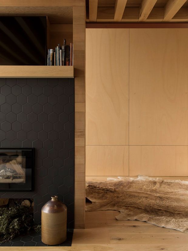 Close-up interior view of a warm timber-lined living area featuring a built-in fireplace with dark hexagonal tile surround, open shelving with books, timber cabinetry, exposed ceiling beams, and a built-in bench or ledge draped with a neutral hide.