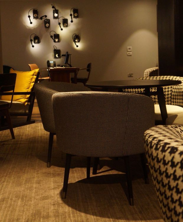 A warm, dimly lit interior lounge or living room furnished with patterned upholstered armchairs and a sofa, a low dark coffee table, and a small work desk in the background. The room features a striking cluster of black wall-mounted light fixtures, neutral taupe walls, and a textured brown floor covering. Bold black-and-white geometric upholstery adds visual contrast.