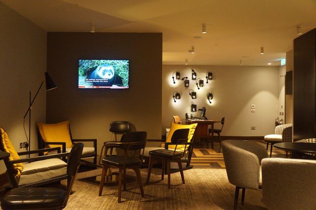 A dimly lit contemporary lounge or media room with multiple lounge chairs and small tables arranged on a textured carpet. A wall-mounted television, decorative wall lighting, and a compact dining or work table are visible. The room uses warm ambient lighting, dark neutral walls, timber furniture, and mustard-yellow upholstered accents.