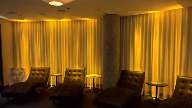 A dimly lit lounge or relaxation area with several tufted leather chaise lounges, small round side tables, full-height golden curtains, recessed ceiling lighting, and a central exposed concrete column. The warm amber lighting creates an intimate, luxurious atmosphere.