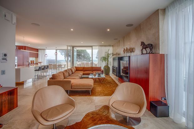 Open-plan mid-century-inspired living room with a warm caramel sectional, sculptural lounge chairs, a large organic-patterned rug, timber cabinetry, and a stone-textured feature wall. The space opens to a dining area and kitchen, with expansive glazing and sheer curtains providing abundant natural light.