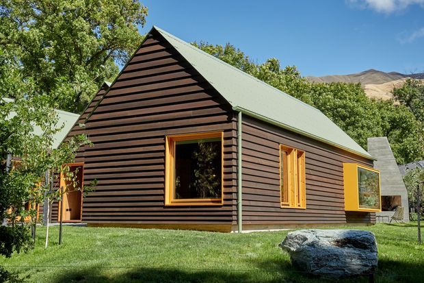 Exterior view of a compact single-storey gable-roof house set in a landscaped garden. The building has dark horizontal timber cladding, a pale green metal pitched roof, warm timber-framed windows and doors, and a prominent angular concrete chimney or fireplace element. Mountains and mature trees are visible in the background.
